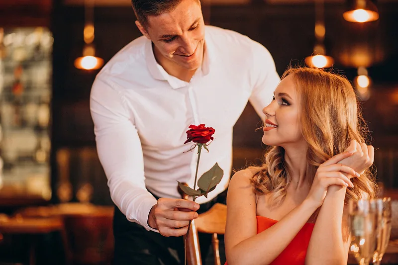 A man bringing a rose to a woman at a restaurant table